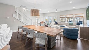 Un vaste espace de vie comprenant une table à manger en bois, des chaises en osier, un pouf bleu, un canapé blanc et un escalier.