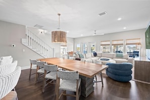 A spacious living area with a wooden dining table, wicker chairs, a blue ottoman, a white sofa, and a staircase. - Between Tides-Pool- Walk to Beach (Murrells Inlet)