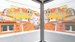 Blick durch ein Fenster auf ein Gebäude mit mehreren Balkonen, orangefarbenen Ziegeldächern und einer Mischung aus gelben und rosa Fassaden.