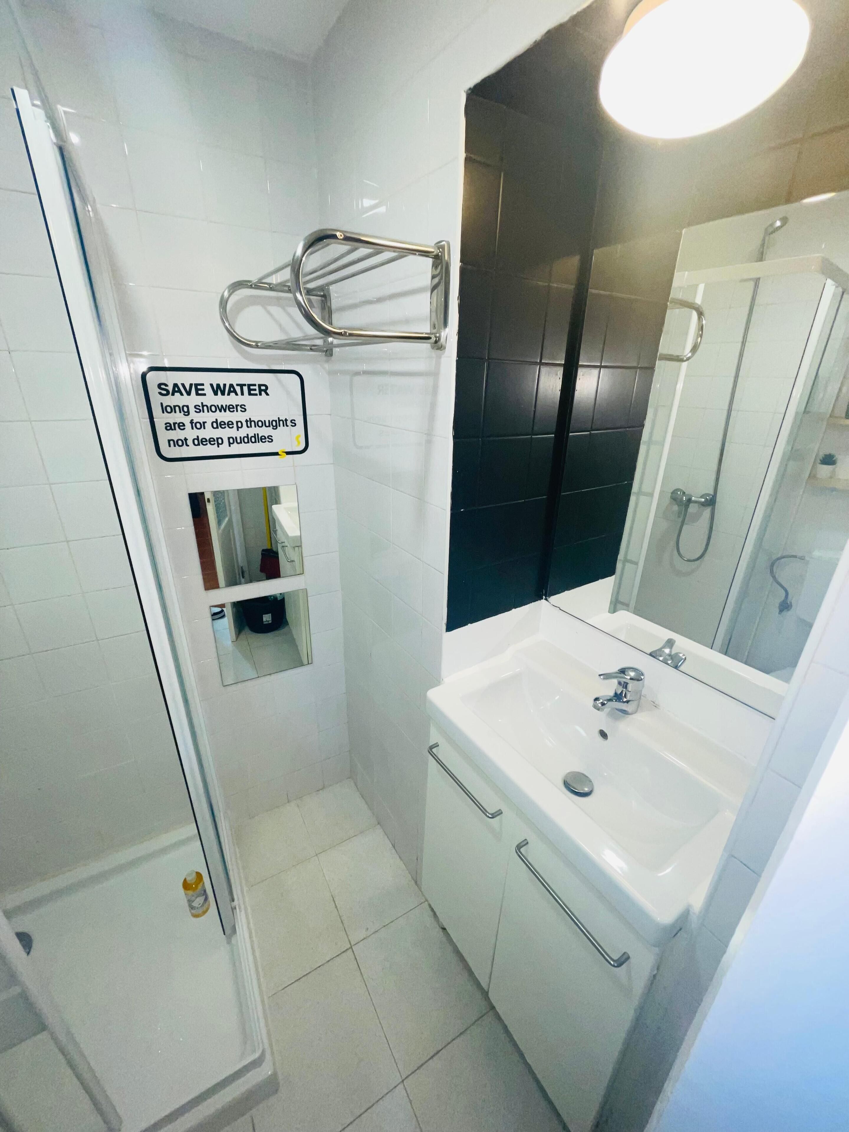 Een moderne badkamer met een glazen douchecabine, een witte wastafel met spiegel, en een bordje dat waterbesparing stimuleert.