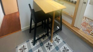 Ein gemütlicher Raum mit einem Holztisch und Stühlen, einem Fliesenboden mit geometrischem Muster, einer Deckenleuchte und einem Regal mit Tassen und Gläsern.