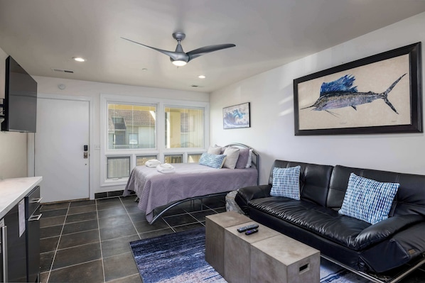 A modern bedroom with a bed, a black leather sofa, a ceiling fan, and artwork on the walls.