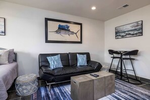 A modern living room with a black leather sofa, a small coffee table, a round ottoman, a bar stool, and two framed artworks on the walls.