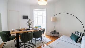 A modern living room with a sofa, a dining table with chairs, a TV mounted on the wall, and a lamp.
