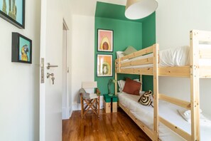 Quarto com beliches, escrivaninha e uma cadeira. Há quadros nas paredes e uma porta com puxador.