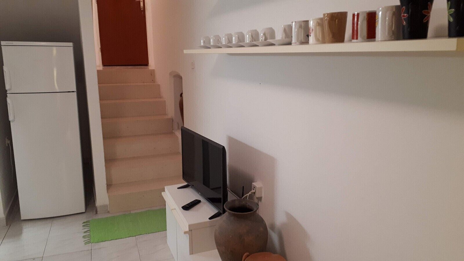A kitchen with a white refrigerator, a TV on a white stand, a vase, and a shelf with various mugs.