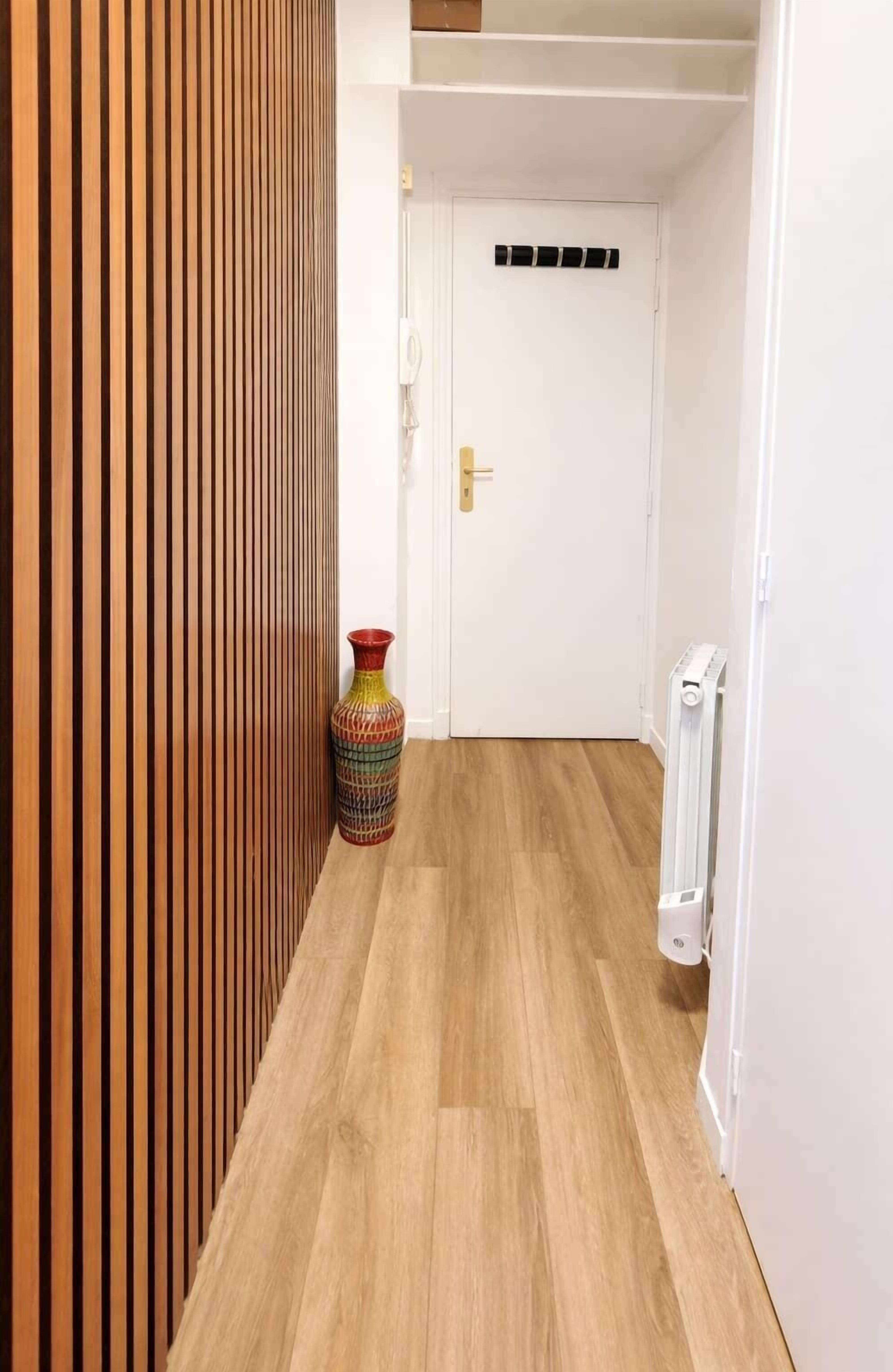Un couloir avec un mur recouvert de lambris en bois, un vase coloré et une porte blanche.