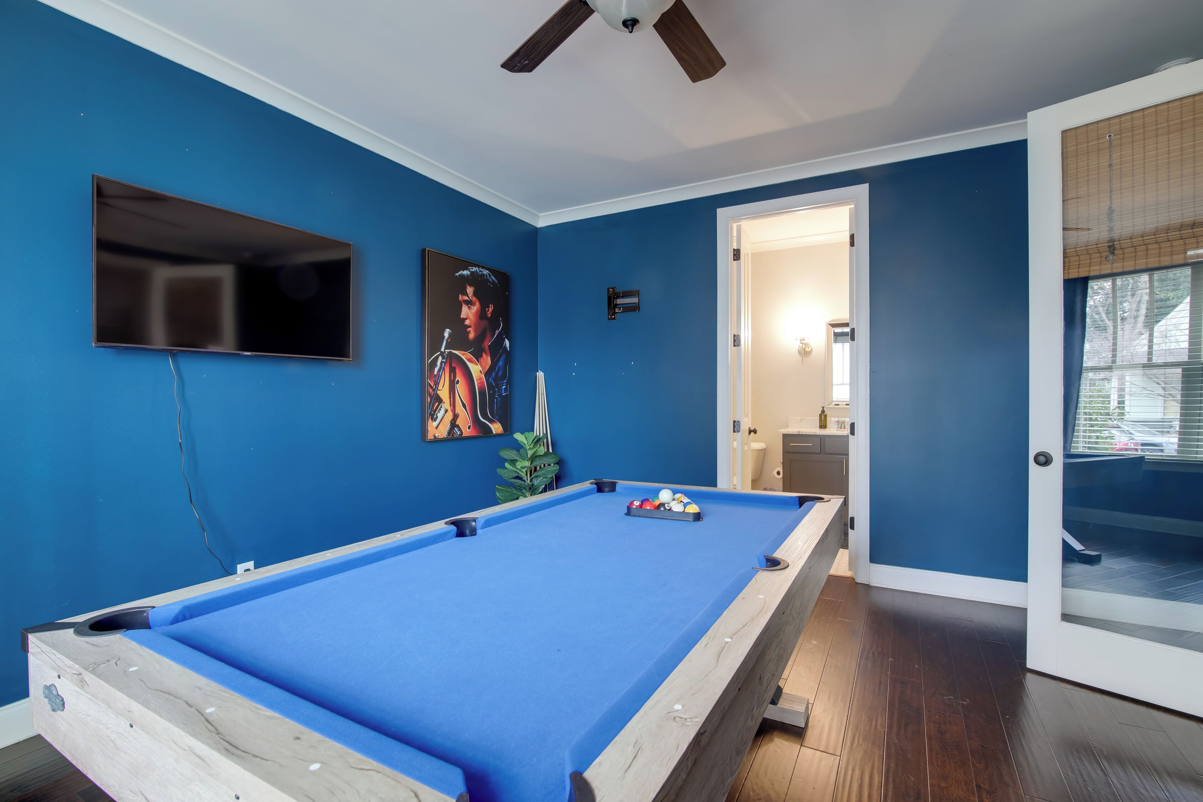 A room with a pool table, a ceiling fan, a door leading to another room, and a wall-mounted TV.