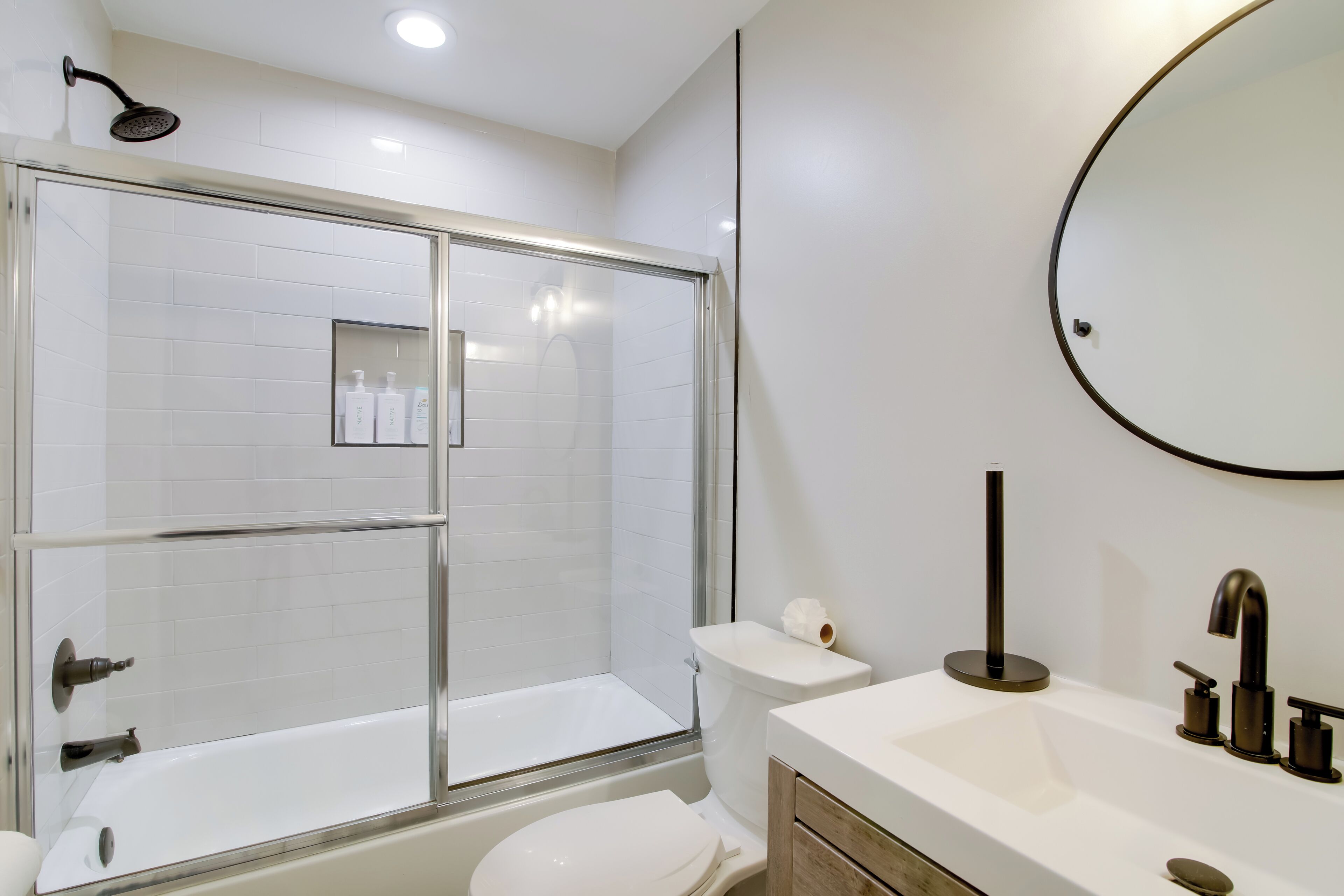 Un bany modern amb dutxa d'acer vitrulat, vàter blanc i un lavabo amb mirall rodó.