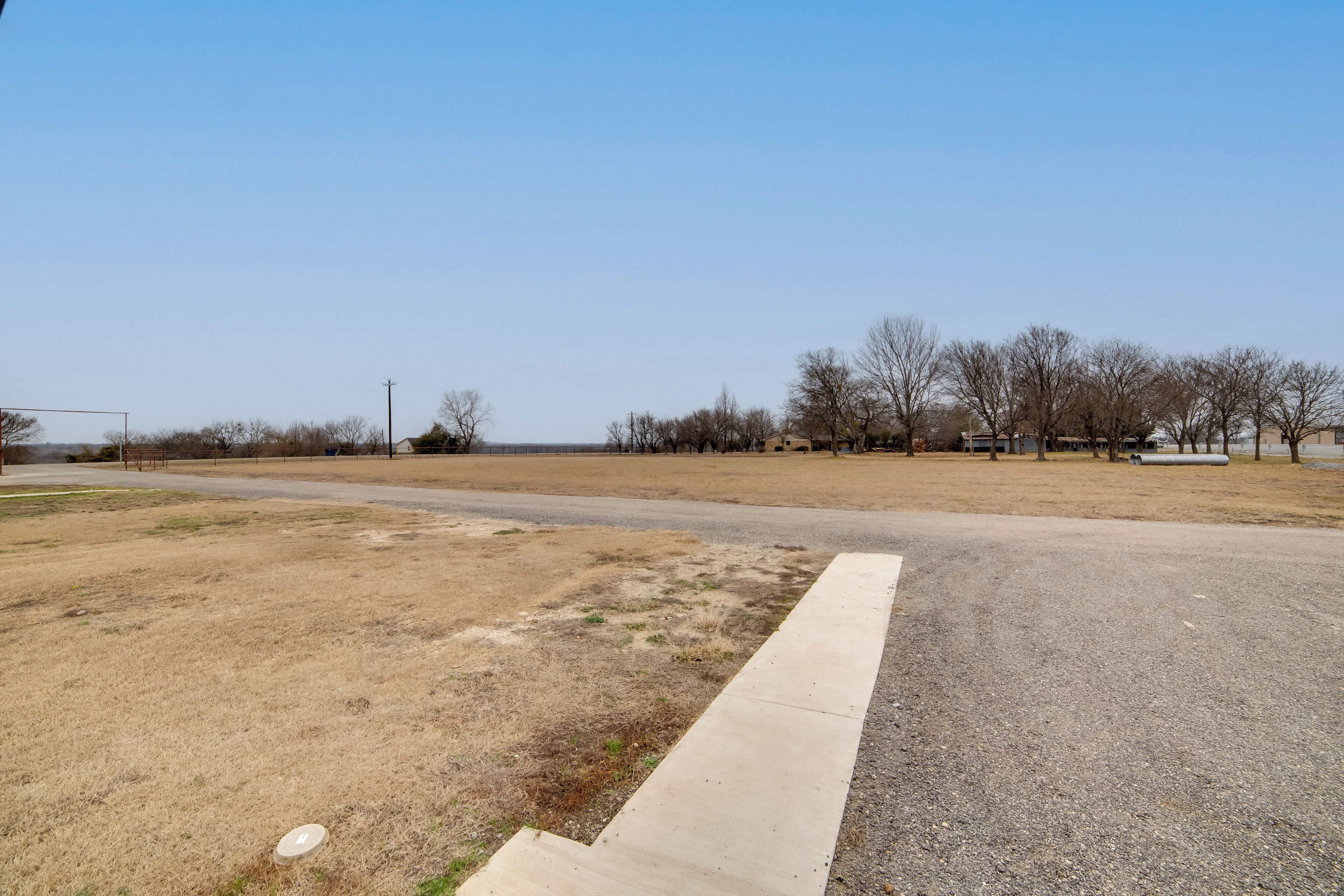 A paved area with a grassy field and trees in the background.