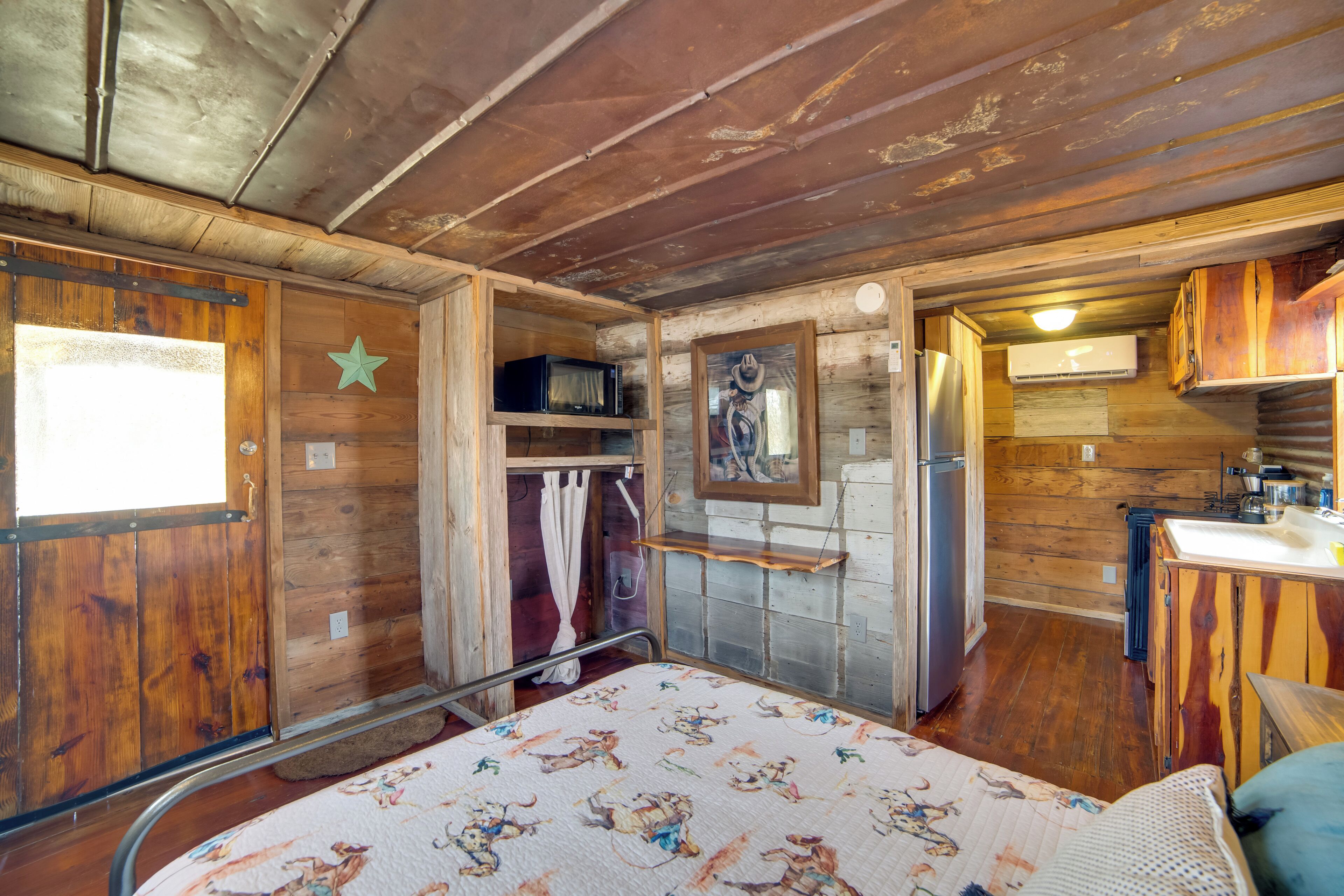 Interior rústico de una cabaña con techo de madera, una cama con colcha floral, una pequeña cocina y un baño con cortina de ducha.