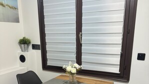 A dining area with a wooden table, two black chairs, a vase with white flowers, and a window with blinds. - Cosy apartment with terrace (Algolsheim)