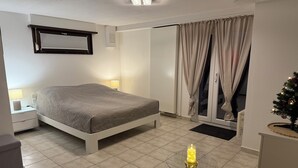 Een slaapkamer met een bed, een nachtkastje, een lamp, een klein tafeltje met een kaars, een rieten stoel en een schuifpui met gordijnen.