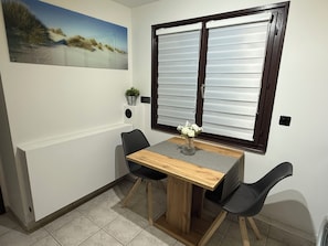 Comedor con mesa de madera, dos sillas negras, un florero con flores, una planta en maceta y una ventana con persianas.