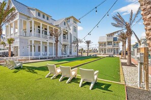 A courtyard with a green lawn, white chairs, and a pool table. - Red White and Saguaro 246 DEW (Port Aransas)