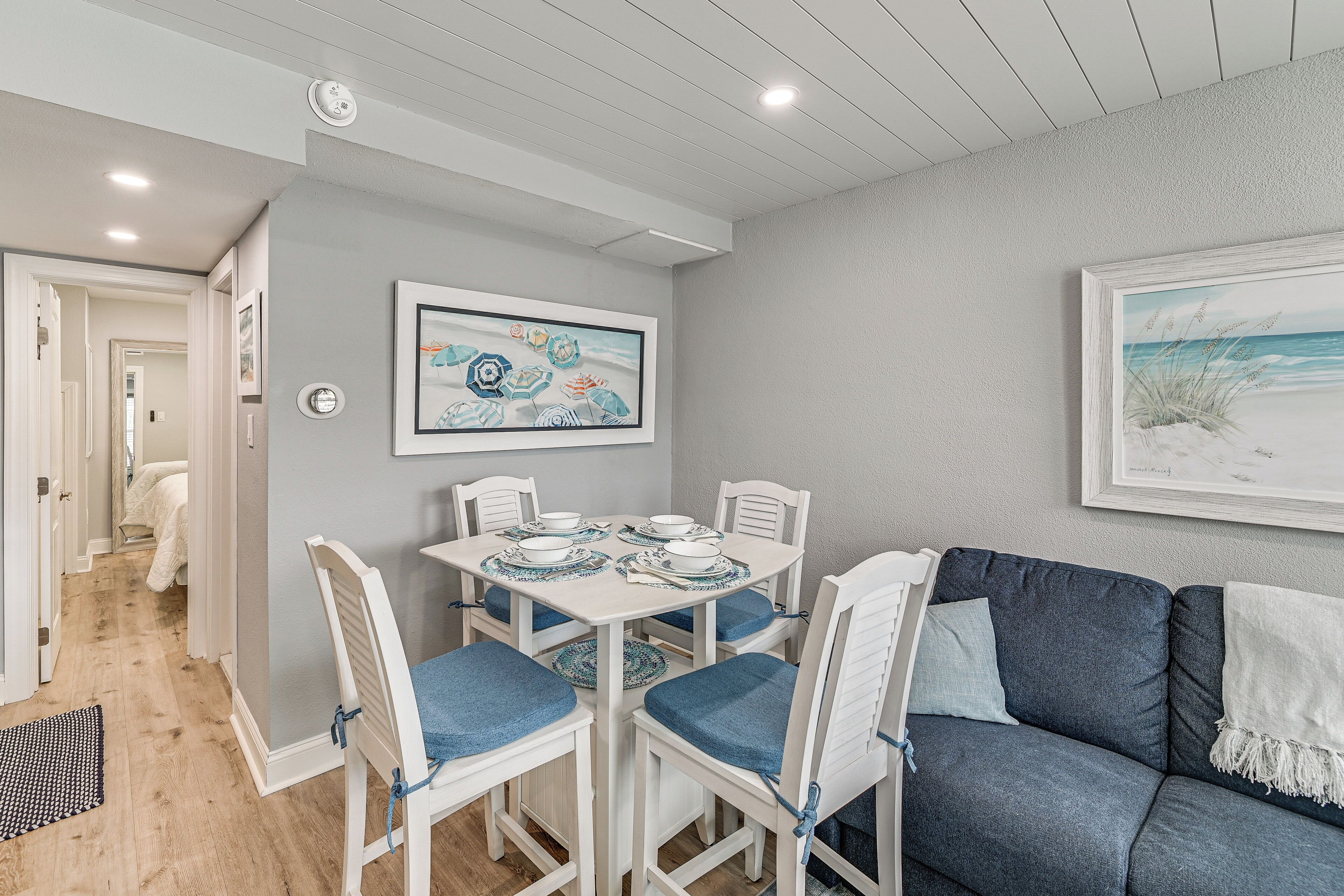 A dining area with a round table, white chairs, and blue cushions. There are framed artworks on the walls and a dark blue sofa with a white throw blanket.