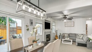 Un vaste salon doté d’une cheminée, un coin repas avec une table et des chaises en bois, un canapé gris et un ventilateur de plafond.