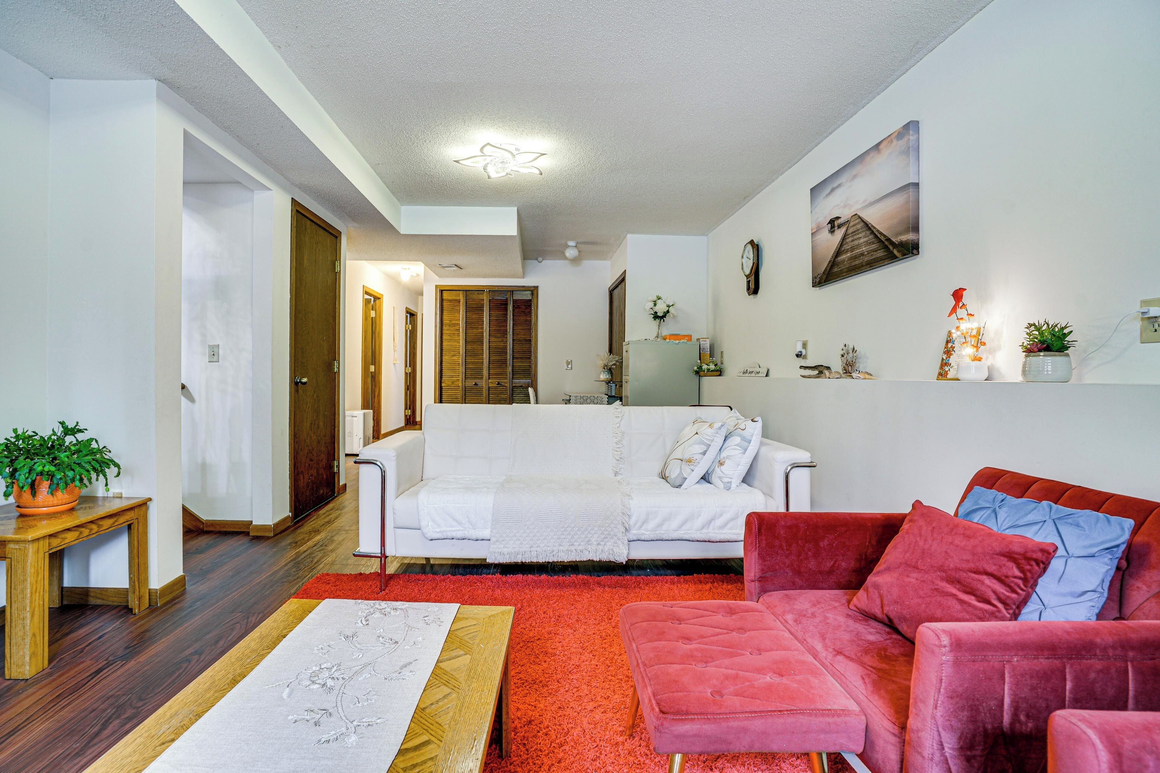A living room with a white sofa, a red sofa, a wooden coffee table, and a red rug. There is a wooden door and a white wall with a painting.