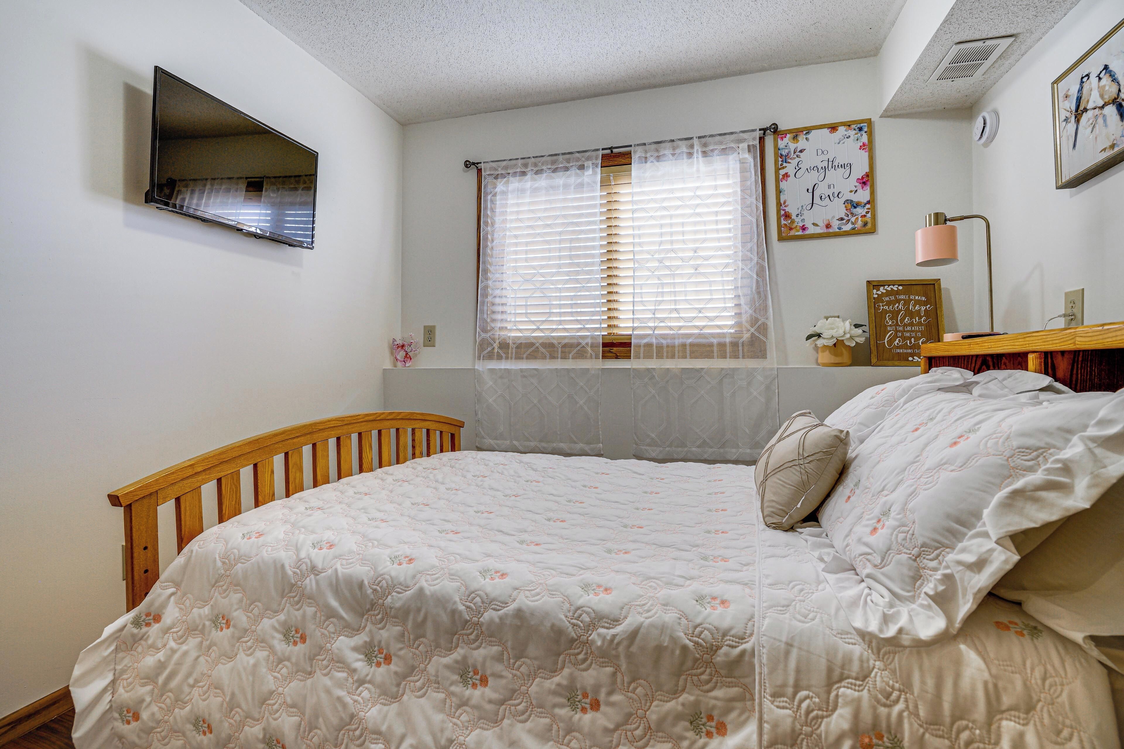 A bedroom with a wooden bed, a window with blinds, a wall-mounted TV, and a framed quote on the wall.