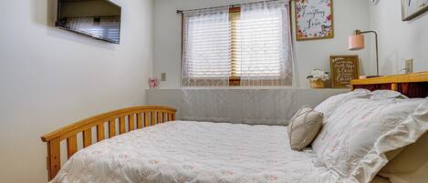A bedroom with a wooden bed, a window with blinds, a wall-mounted TV, and a framed quote on the wall.