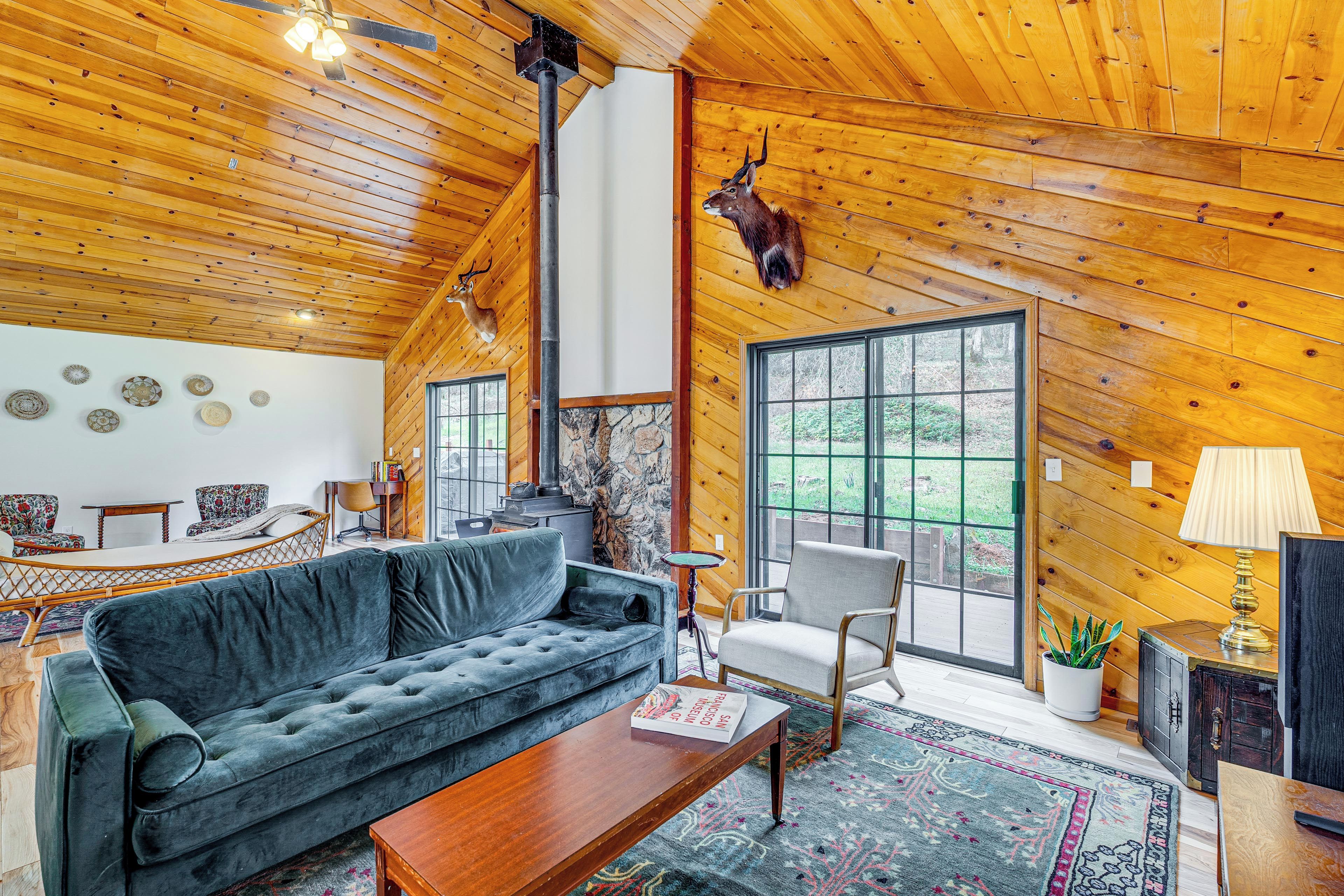 A cozy cabin interior with a wooden ceiling, a fireplace, a sofa, a chair, and a deer head mounted on the wall.