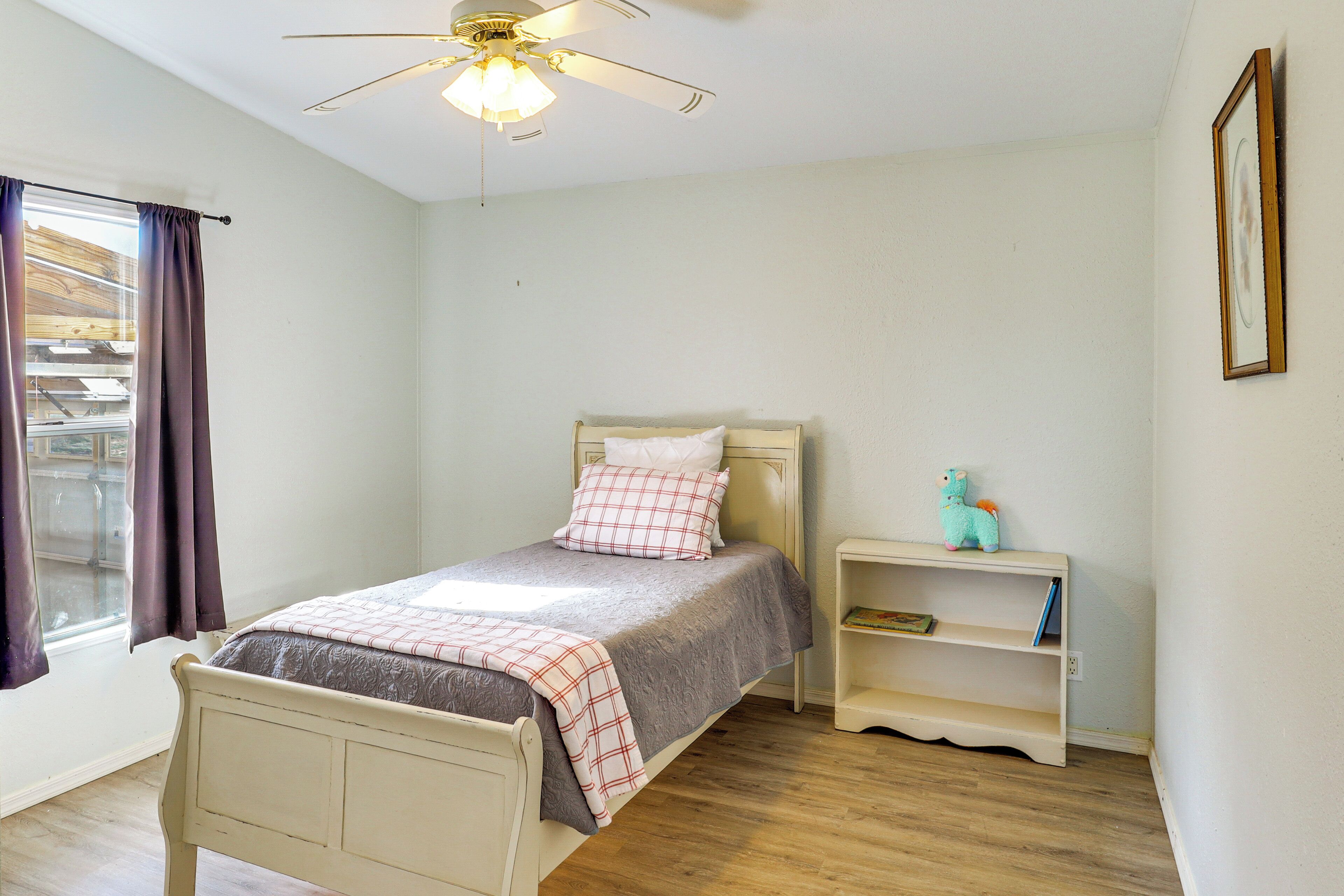 Une chambre à coucher comprenant un lit, une table de chevet, un ventilateur de plafond, une fenêtre avec des rideaux et une bibliothèque.