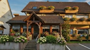 Ein zweistöckiges Gebäude mit einem geneigten Dach, hölzernen Balkonen mit Blumenkästen und einem Eingangsbereich mit Stufen.
