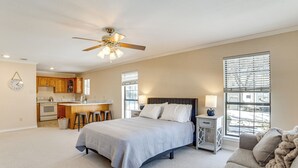 A bedroom with a bed, a nightstand, a lamp, a window with blinds, a ceiling fan, and a kitchen area in the background. - 'texoma Shores Hideaway' Family Home w/ Water View (Pottsboro)