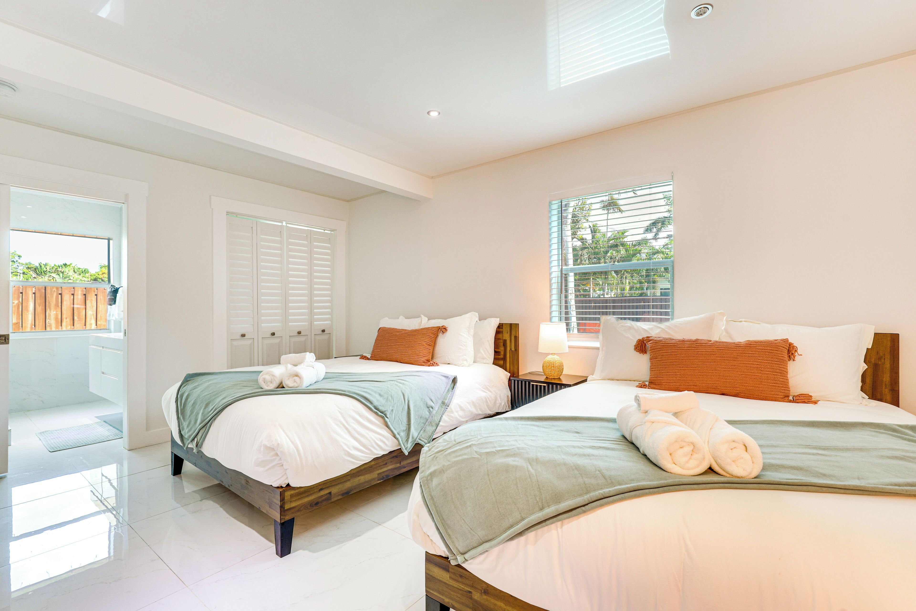 A hotel room with two beds, a window with blinds, and a view of greenery outside.