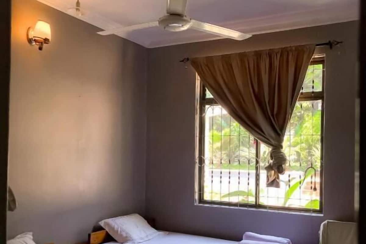 A room with two single beds, a ceiling fan, a window with curtains, and a chair.