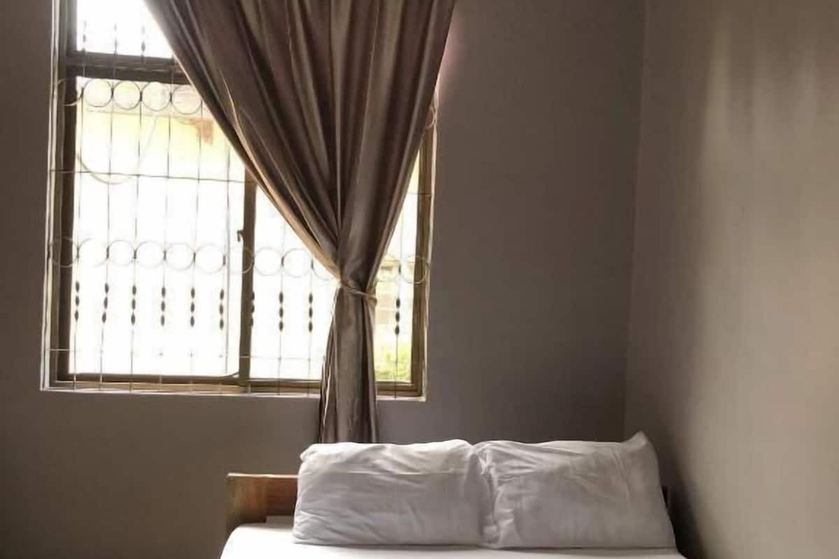 A single bed with white linens, a window with a curtain, and a ceiling fan.