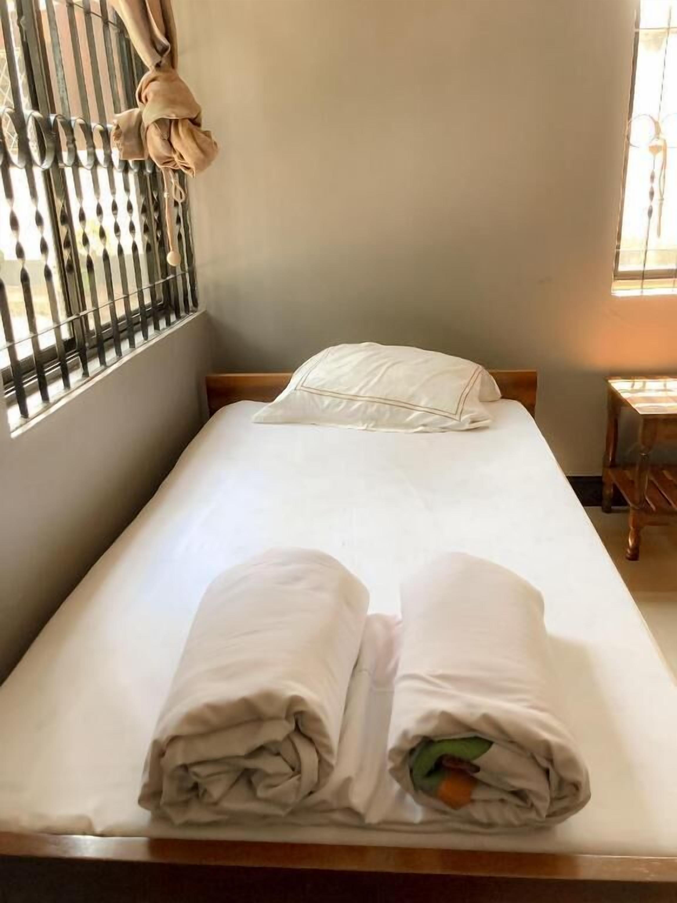 A single bed with white linens, a wooden headboard, and a window with bars and a curtain.
