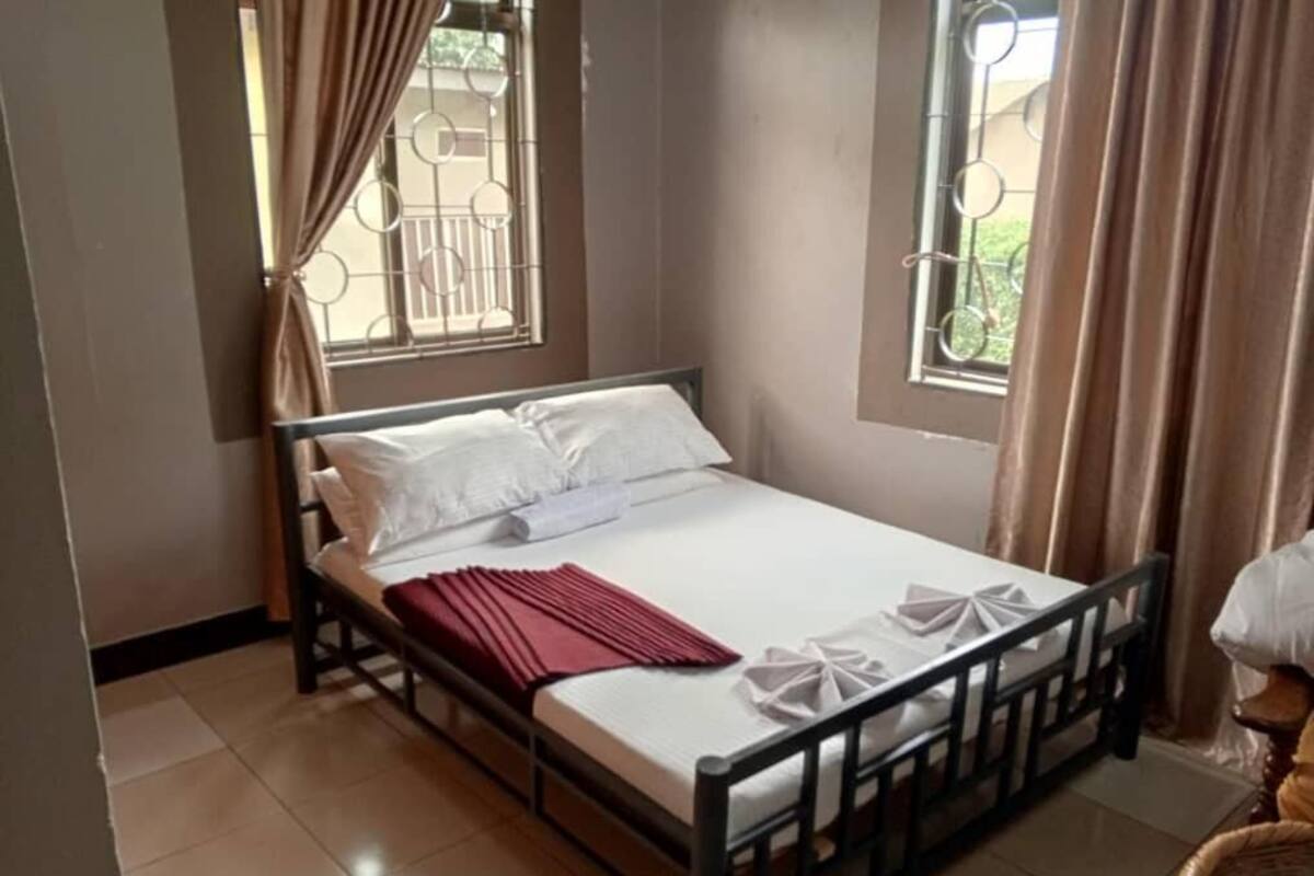 A single bed with a metal frame, white bedding, and a red blanket. There are two windows with curtains, one on each side of the bed.