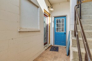 Un pasillo estrecho con una puerta azul, un escalón pequeño y una barandilla de metal que conduce a una escalera.