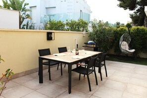 Een terras buiten met een tafel voor vier personen, omringd door stoelen, een barbecue en een hangstoel.