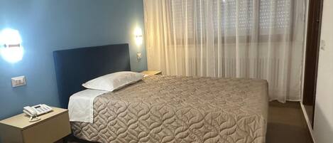 Una habitación de hotel con una cama, una mesita de noche con un teléfono y una ventana con cortinas traslúcidas.