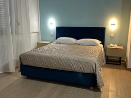 Una cama bien tendida con un cabecero azul, dos mesitas de noche con lámparas y una ventana con cortinas.