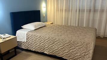 Quarto de hotel com cama, mesa de cabeceira com telefone, criadeira e janela com cortinas leves.