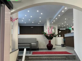 Zona de recepción con mostrador, un sofá gris, alfombra roja y un jarrón grande con flores rosadas.