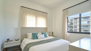 Ett hotellrum med en säng, ett skrivbord, ett fönster med gardiner och utsikt över byggnader.