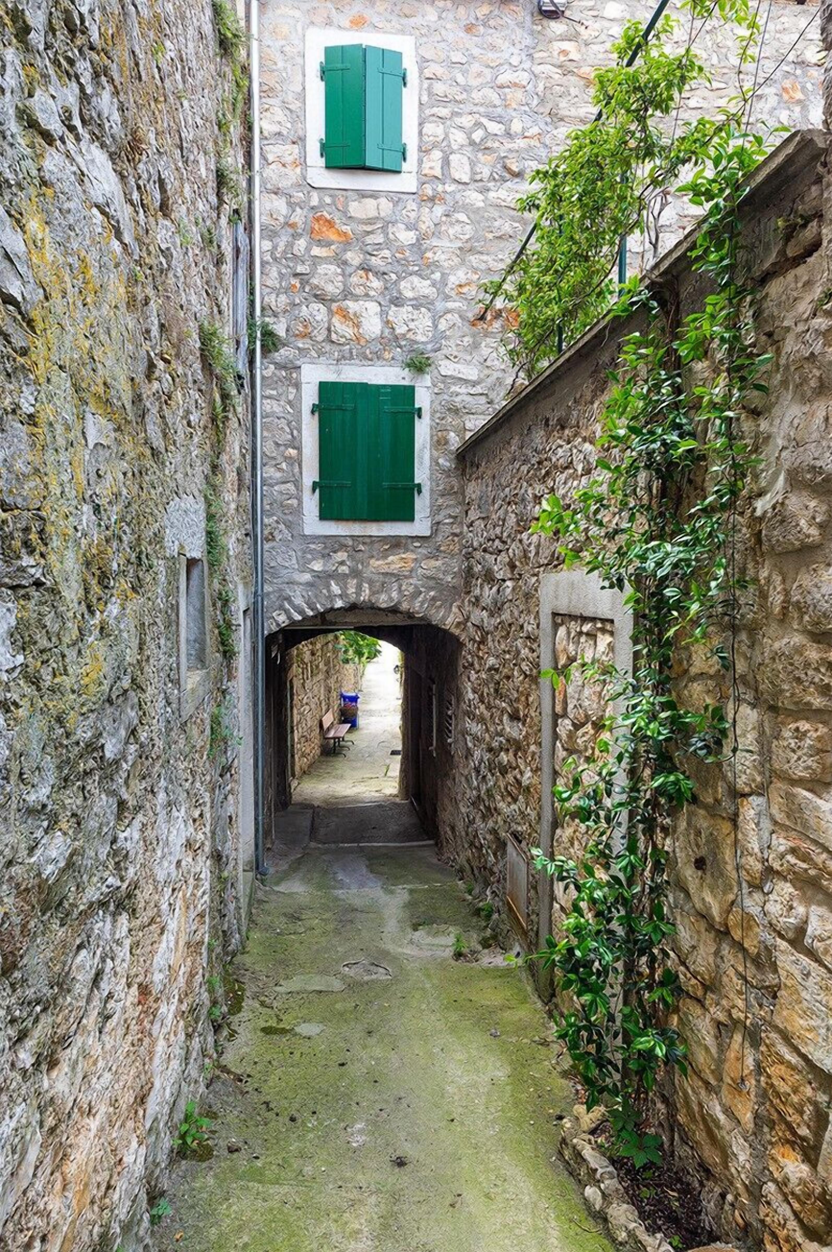 A narrow stone alleyway with an archway leading to a building with green shutters.