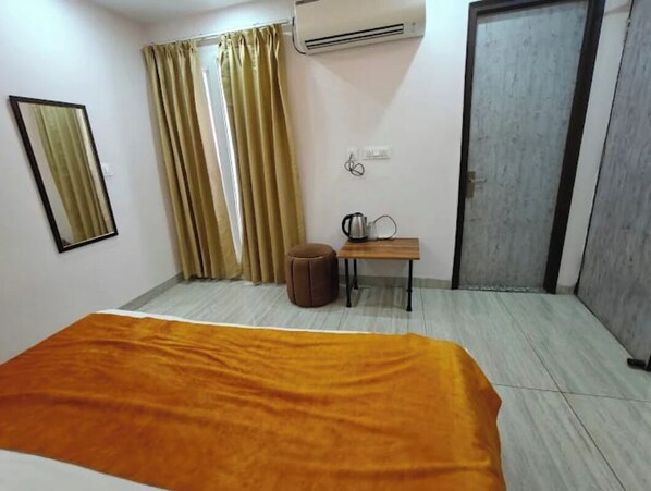 A hotel room with a bed, a small table with a kettle, a brown ottoman, a mirror, and an air conditioning unit. - Yog Namah Hotel & Cafe (Rishikesh)