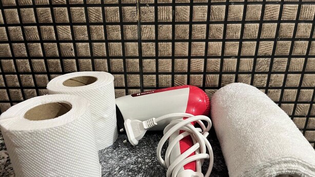 A bathroom with a tiled wall, two rolls of toilet paper, a red and white hairdryer, and a rolled-up towel. - Yog Namah Hotel & Cafe (Rishikesh)