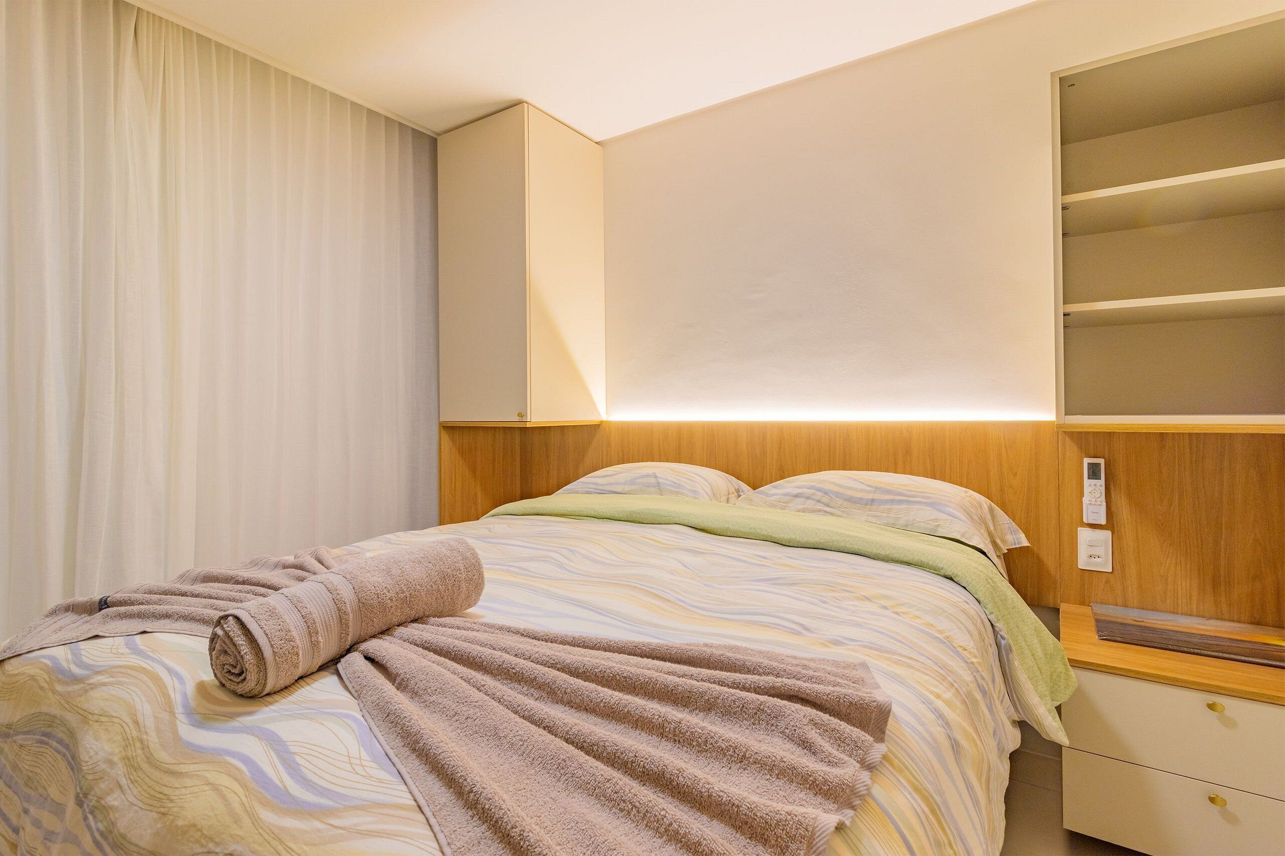 A hotel room with a bed, a nightstand, a wall-mounted shelf, and a window with curtains.