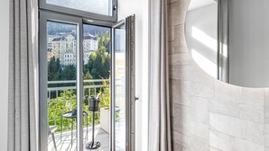 A modern interior with a sliding glass door leading to a balcony, a round mirror, and a light-colored tiled wall. - Spacious XXL Deluxe suite with private balcony and village views. (Bad Gastein)