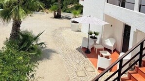Un edificio de dos pisos con fachada blanca y tejado de tejas rojas, un espacio al aire libre cubierto con sillas y mesa blancas, y una palmera en primer plano.