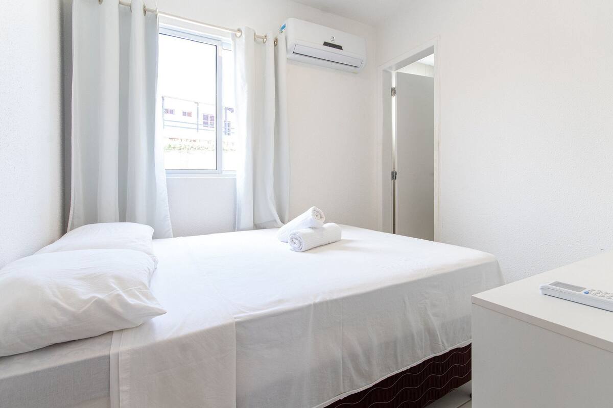 Quarto de hotel com cama, janela com cortinas, ar condicionado, mesa de cabeceira e porta.