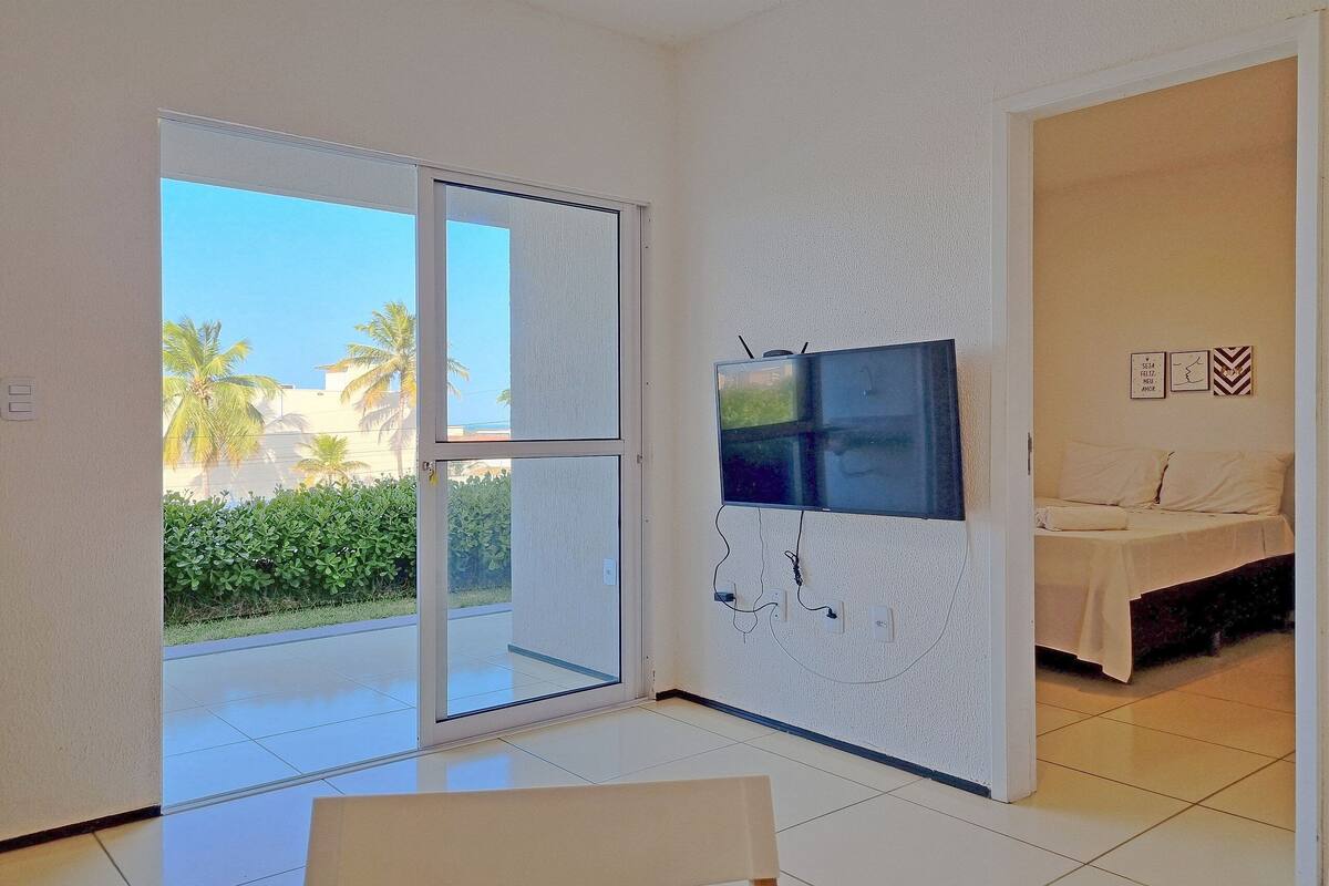 Quarto com uma cama, televisão na parede e porta de vidro deslizante para área externa com palmeiras.