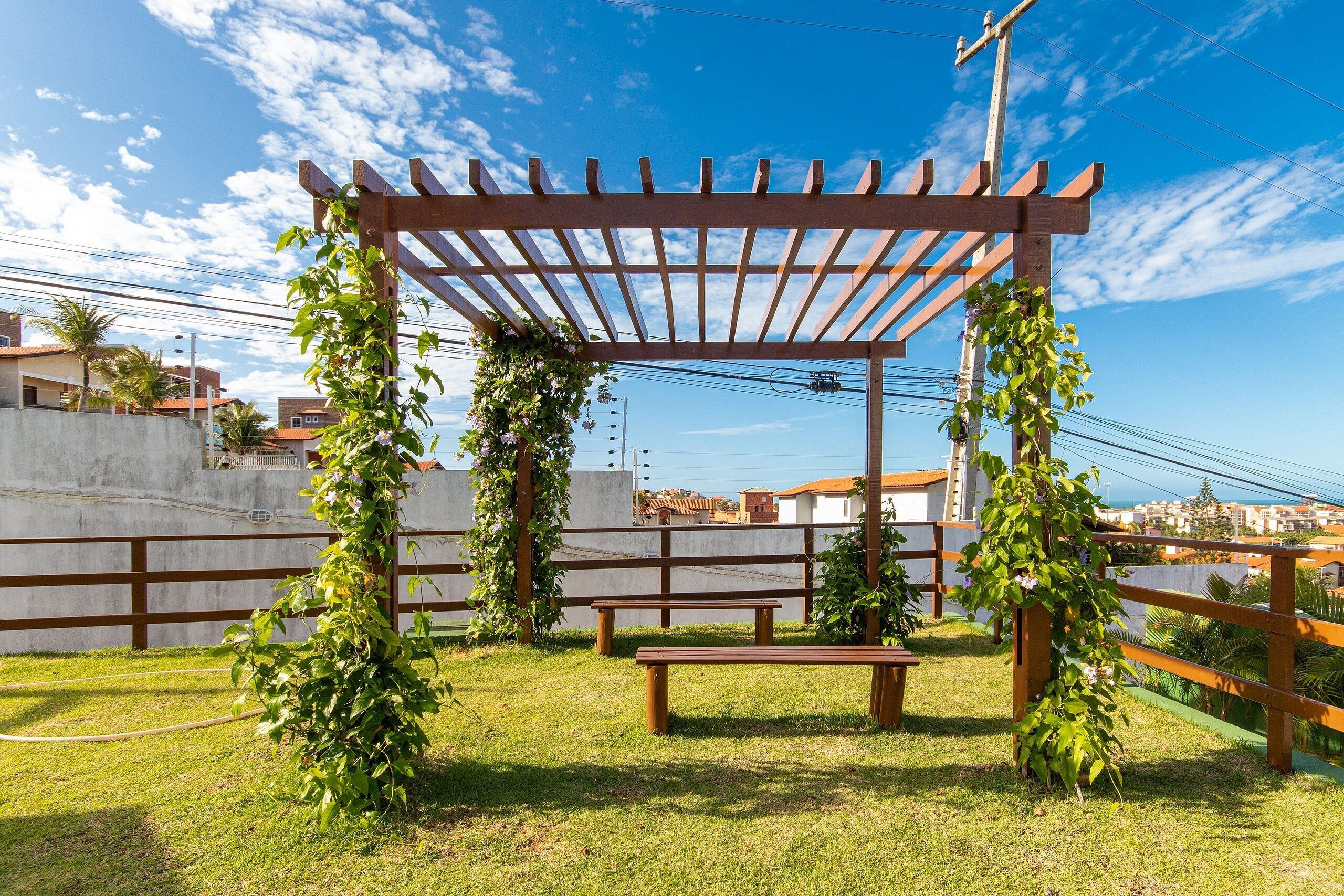 Una pérgola de madera con un banco, rodeada de vegetación y un cielo despejado.