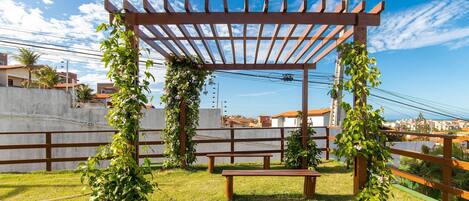 Una pérgola de madera con un banco, rodeada de vegetación y un cielo despejado.